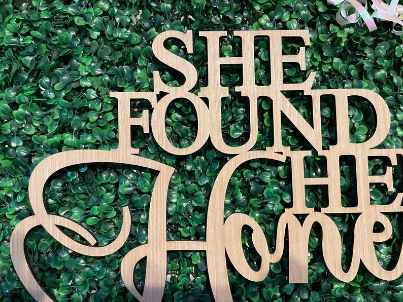 Peut inclure: Panneau en bois avec l'inscription "SHE FOUND HER HOME" dans une police d&eacute;corative. Le panneau est en bois de couleur claire et est plac&eacute; sur un fond de feuillage vert.