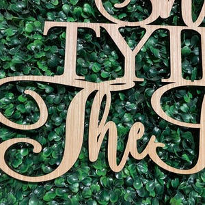 Puede incluir: Letrero de madera con las palabras "TYING the Knot" en fuente cursiva. El letrero est&aacute; hecho de madera de color claro y est&aacute; colocado sobre un fondo de follaje verde. Una cinta rosa es visible en la esquina superior izquierda.
