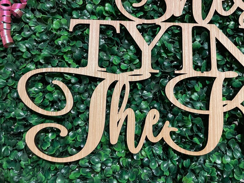 Puede incluir: Letrero de madera con las palabras "TYING the Knot" en fuente cursiva. El letrero est&aacute; hecho de madera de color claro y est&aacute; colocado sobre un fondo de follaje verde. Una cinta rosa es visible en la esquina superior izquierda.
