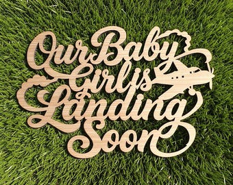 May include: A light brown wooden cutout sign featuring the cursive text Our Baby Girl is Landing Soon with an integrated graphic of a small propeller airplane flying through clouds, showcasing a natural wood grain texture and laser-cut edges.