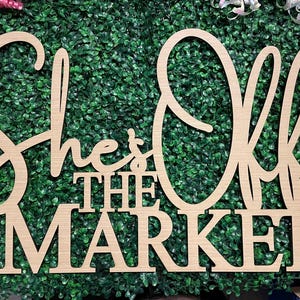 May include: A wooden sign with the words "She's Off The Market" in a decorative font. The sign is made of light-colored wood and is set against a backdrop of green foliage. The sign is likely used for wedding or engagement announcements.