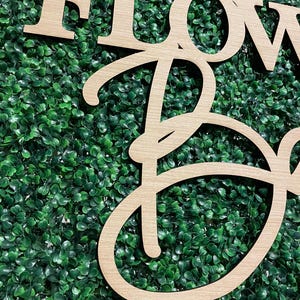 May include: Wooden sign with the words "FLOWER" and "BOX" in a cursive font. The sign is set against a backdrop of green artificial foliage.