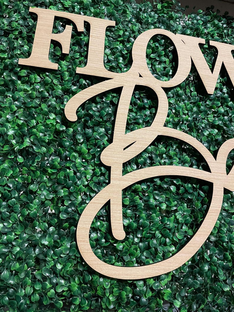 May include: Wooden sign with the words "FLOWER" and "BOX" in a cursive font. The sign is set against a backdrop of green artificial foliage.