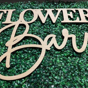 May include: Wooden sign with the words "Flower Bar" in a cursive font. The sign is made of light brown wood and is sitting on a green artificial grass background.