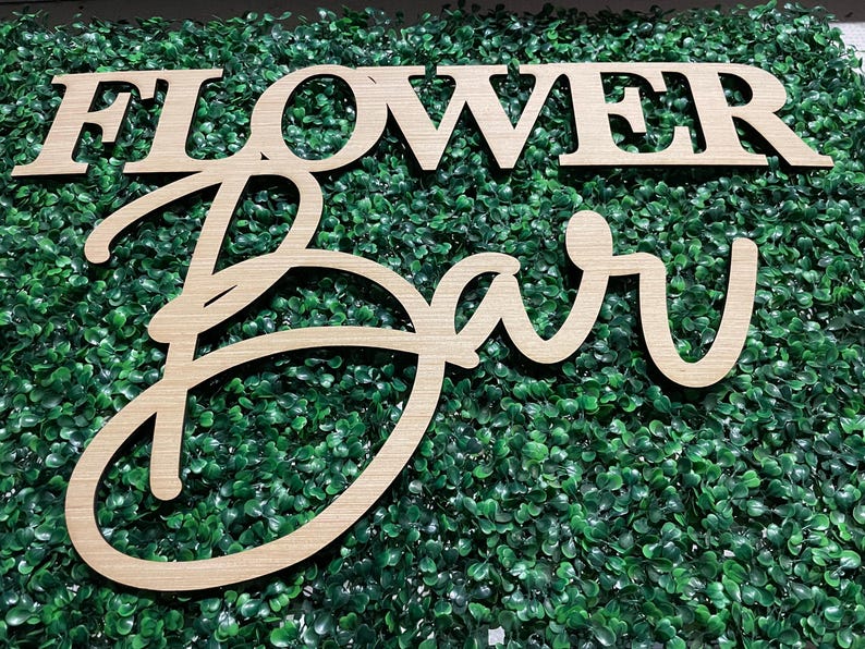 May include: Wooden sign with the words "Flower Bar" in a cursive font. The sign is made of light brown wood and is sitting on a green artificial grass background.