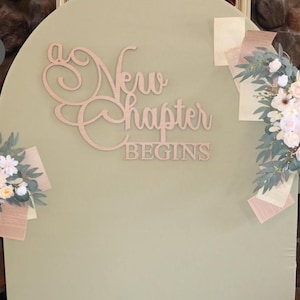 May include: A light green arched backdrop with the words "a New chapter BEGINS" in wooden script. Floral arrangements with white and pink flowers and greenery are attached to the backdrop.