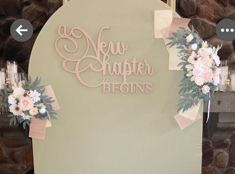 May include: A light green arched backdrop with the words "a New chapter BEGINS" in wooden script. Floral arrangements with white and pink flowers and greenery are attached to the backdrop.
