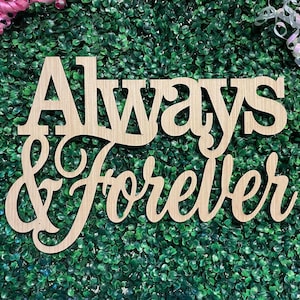 May include: A light-colored wooden cutout sign featuring the words Always & Forever in a combination of serif and cursive script fonts. The sign is positioned against a textured green background with pink and white ribbon accents in the corners.
