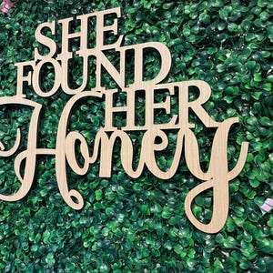 Peut inclure: Panneau en bois avec l'inscription "SHE FOUND HER HONEY" dans une police d&eacute;corative. Le panneau est en bois de couleur claire et est plac&eacute; sur un fond de feuillage vert. Des rubans sont visibles en arri&egrave;re-plan.