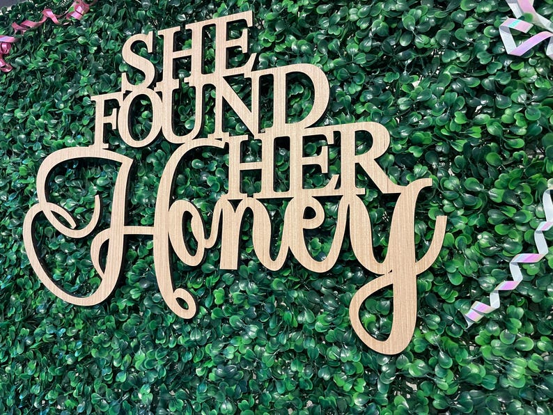 Peut inclure: Panneau en bois avec l'inscription "SHE FOUND HER HONEY" dans une police d&eacute;corative. Le panneau est en bois de couleur claire et est plac&eacute; sur un fond de feuillage vert. Des rubans sont visibles en arri&egrave;re-plan.
