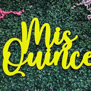 Op de afbeelding: Een felgeel bord met de tekst Mis Quince in een cursief lettertype. Het bord is geplaatst tegen een donkergroene bladerrijke achtergrond met roze krullende linten in de linker- en rechterbovenhoek van de weergave.