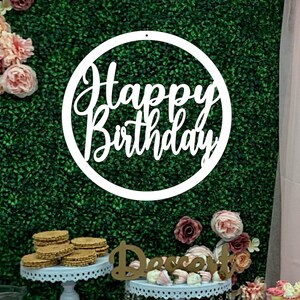 May include: White wooden circle sign with the text "Happy Birthday" in a cursive font. The sign is hanging on a green artificial grass backdrop with pink flowers. There are desserts on a table in the foreground.
