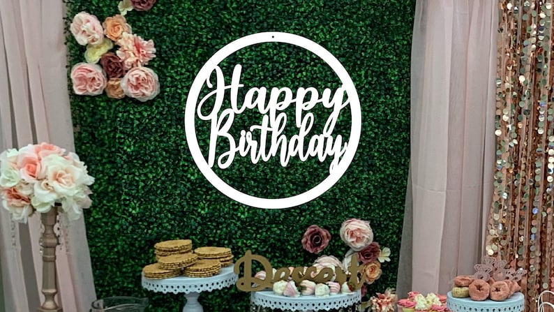 May include: White wooden circle sign with the text "Happy Birthday" in a cursive font. The sign is hanging on a green artificial grass backdrop with pink flowers. There are desserts on a table in the foreground.