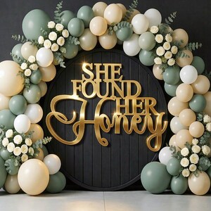 K&ouml;nnte beinhalten: Ein runder schwarzer Holzhintergrund mit dem goldenen Schriftzug SHE FOUND HER HONEY, umrahmt von einem Bogen aus salbeigr&uuml;nen und cremefarbenen Luftballons, durchsetzt mit kleinen wei&szlig;en Bl&uuml;ten und gr&uuml;nen Eukalyptusbl&auml;ttern.