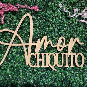 May include: Wooden sign with the words "Amor Chiquito" in a cursive and block font. The sign is made of light-colored wood and is set against a green background with pink and white ribbons.