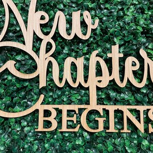 May include: Wooden sign with the words "New Chapter Begins" in a cursive font. The sign is made of light-colored wood and is set against a backdrop of green foliage. The sign is a decorative item.
