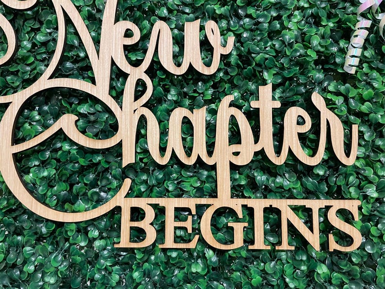 May include: Wooden sign with the words "New Chapter Begins" in a cursive font. The sign is made of light-colored wood and is set against a backdrop of green foliage. The sign is a decorative item.