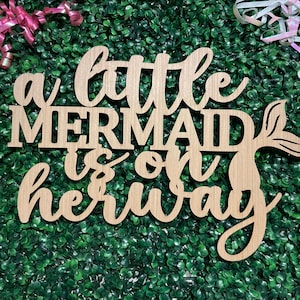 May include: Wooden sign with the phrase "a little MERMAID is on her way" in a cursive font. The sign has a mermaid tail design and is set against a green background with pink and white ribbons.