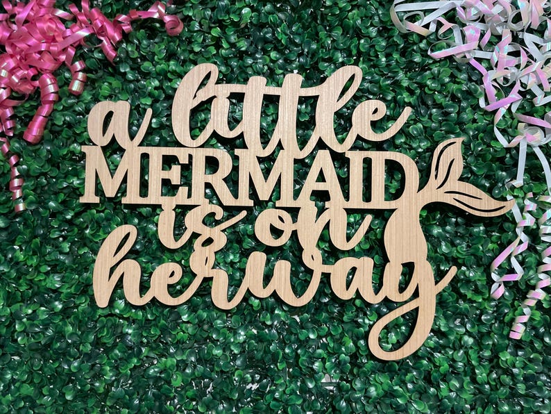 May include: Wooden sign with the phrase "a little MERMAID is on her way" in a cursive font. The sign has a mermaid tail design and is set against a green background with pink and white ribbons.