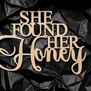 K&ouml;nnte beinhalten: Ein hellbraunes Holzschild mit dem Schriftzug SHE FOUND HER HONEY in einer Kombination aus Block- und Schreibschrift. Das Schild ist aus einem flachen, glatten Material mit nat&uuml;rlicher Holzmaserung und sauberen, lasergeschnittenen Kanten gefertigt.