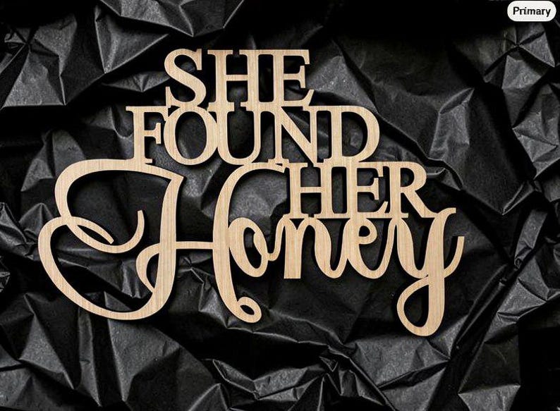 K&ouml;nnte beinhalten: Ein hellbraunes Holzschild mit dem Schriftzug SHE FOUND HER HONEY in einer Kombination aus Block- und Schreibschrift. Das Schild ist aus einem flachen, glatten Material mit nat&uuml;rlicher Holzmaserung und sauberen, lasergeschnittenen Kanten gefertigt.