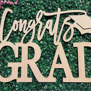 May include: Wooden sign with the words "Congrats Grad" in a cursive font and block letters, with a graduation cap silhouette. The sign is set against a backdrop of green foliage and pink ribbon.