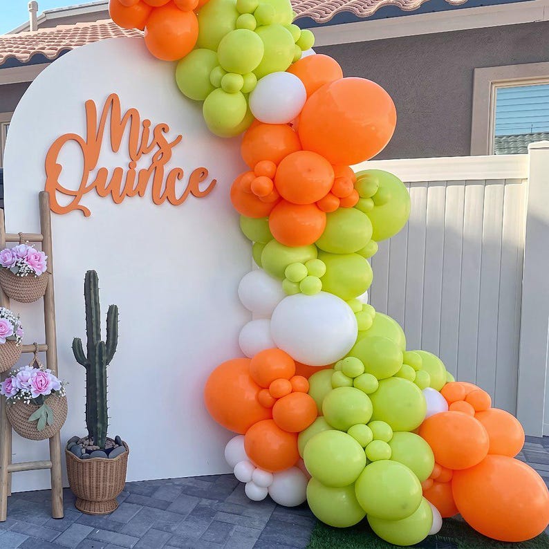 Op de afbeelding: Een witte boogvormige achtergrond met de oranje tekst Mis Quince, versierd met een grote ballonslinger in limoengroen, feloranje en wit. Een houten ladder bevat drie gevlochten manden met roze bloemen en een cactus in een pot.