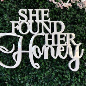 K&ouml;nnte beinhalten: Ein wei&szlig;es, gl&auml;nzendes, dreidimensionales Schild mit dem Text "SHE FOUND HER HONEY" in einer Kombination aus fetter Blockschrift und eleganter Schreibschrift. Die Buchstaben sind erhaben und haben eine glatte, reflektierende Oberfl&auml;che.