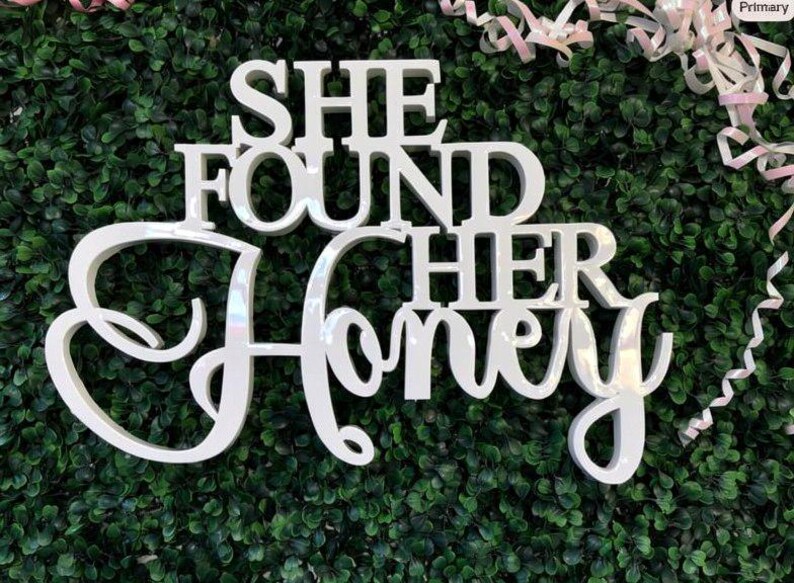 K&ouml;nnte beinhalten: Ein wei&szlig;es, gl&auml;nzendes, dreidimensionales Schild mit dem Text "SHE FOUND HER HONEY" in einer Kombination aus fetter Blockschrift und eleganter Schreibschrift. Die Buchstaben sind erhaben und haben eine glatte, reflektierende Oberfl&auml;che.