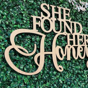 Peut inclure: Panneau en bois clair avec l'inscription "SHE FOUND HER Honey" dans une police d&eacute;corative. Le panneau est plac&eacute; devant un fond de feuillage vert. Un ruban blanc est visible dans le coin sup&eacute;rieur droit.