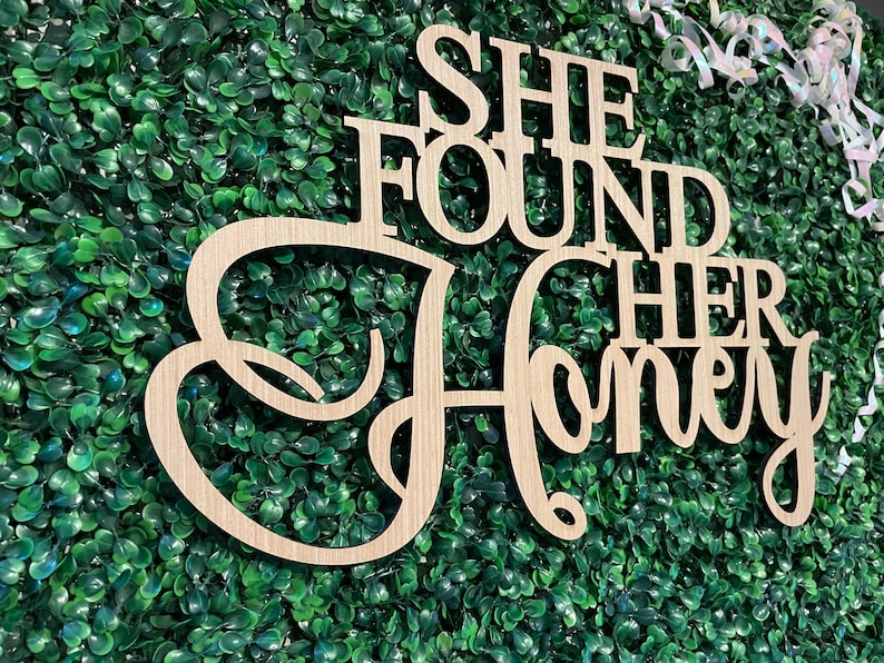 Peut inclure: Panneau en bois clair avec l'inscription "SHE FOUND HER Honey" dans une police d&eacute;corative. Le panneau est plac&eacute; devant un fond de feuillage vert. Un ruban blanc est visible dans le coin sup&eacute;rieur droit.