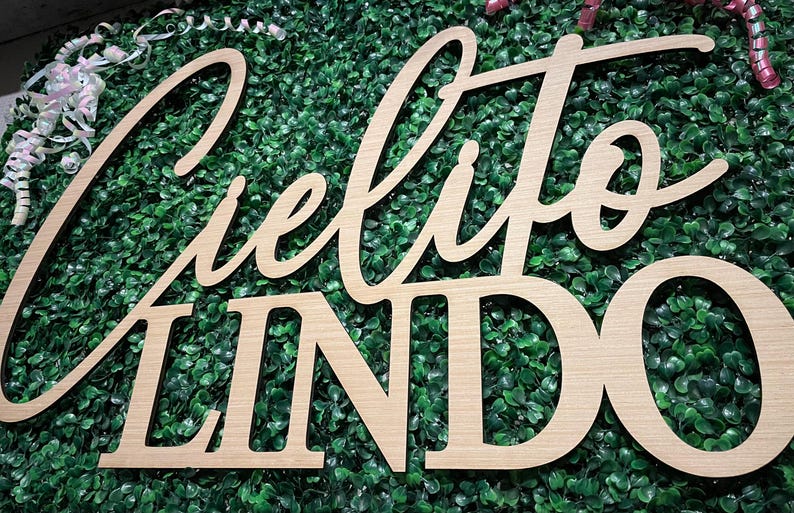May include: Wooden sign with the words "Cielito Lindo" in a cursive and block font. The sign is light brown and set against a backdrop of green foliage. Decorative ribbons are visible in the upper left and right corners.