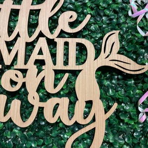 May include: Wooden sign with the words "Little Mermaid on the way" in a cursive font. The sign has a mermaid tail design and is set against a green background with decorative ribbons.