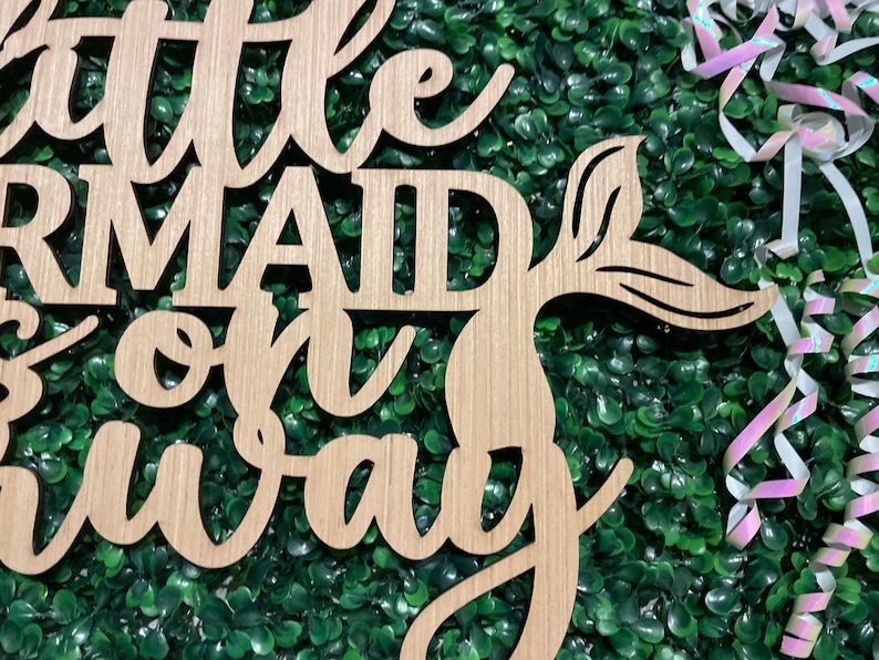 May include: Wooden sign with the words "Little Mermaid on the way" in a cursive font. The sign has a mermaid tail design and is set against a green background with decorative ribbons.