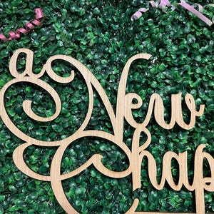 May include: A wooden sign with the words "A New Chapter" in a decorative font. The sign is light brown and set against a backdrop of green foliage and pink and white ribbons.