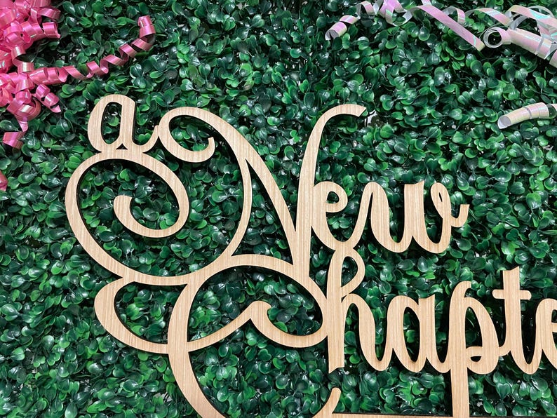 May include: A wooden sign with the words "A New Chapter" in a decorative font. The sign is light brown and set against a backdrop of green foliage and pink and white ribbons.