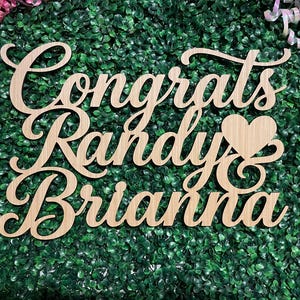 May include: A light-colored wooden cutout sign featuring the text Congrats Randy & Brianna with a heart symbol integrated into the design. The script lettering is connected and smooth, showcasing the natural grain of the wood material.