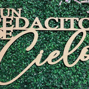 May include: Wooden sign with the phrase "Un Pedacito De Cielo" in a cursive font, meaning "A Little Piece of Heaven". The sign is made of light-colored wood and set against a green textured background.