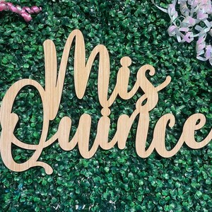 Op de afbeelding: Een lichtbruin houten uitsnede bord met de cursieve tekst Mis Quince. De letters zijn in &eacute;&eacute;n stuk verbonden met een gladde afwerking, waarbij het natuurlijke houtnerfpatroon over het gehele oppervlak van de letters zichtbaar is.