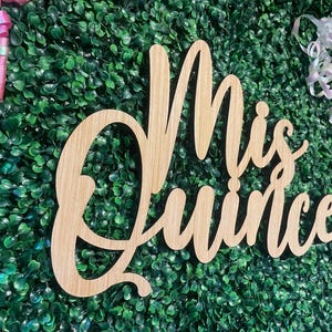 Op de afbeelding: Een lichtgekleurd houten bord met cursieve letters die Mis Quince spellen. Het bord is geplaatst tegen een dichte muur van kunstmatig groen blad en is geaccentueerd met roze en iriserende witte krullende linten op de bovenhoeken.