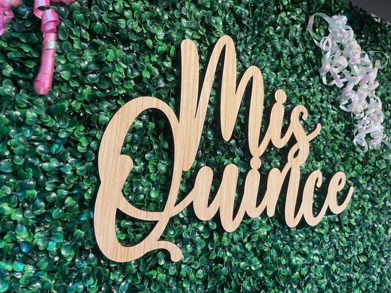 Op de afbeelding: Een lichtgekleurd houten bord met cursieve letters die Mis Quince spellen. Het bord is geplaatst tegen een dichte muur van kunstmatig groen blad en is geaccentueerd met roze en iriserende witte krullende linten op de bovenhoeken.