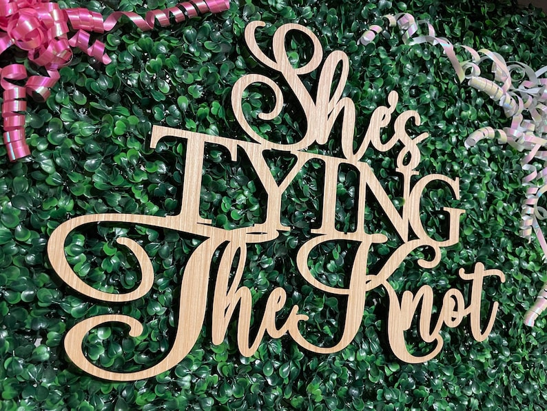 Puede incluir: Letrero de madera con la frase "She's Tying the Knot" en fuente cursiva. El letrero es de madera clara y est&aacute; sobre un fondo de follaje verde. Cintas rosas y blancas en el fondo.