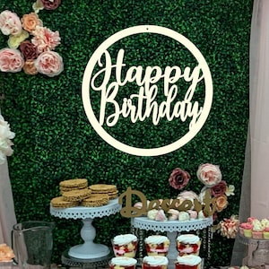 May include: A dessert table decorated with pink and white flowers, a green backdrop, and a wooden circle sign that says "Happy Birthday".