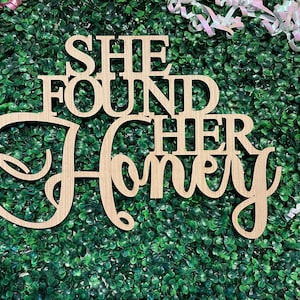 Peut inclure: Panneau en bois clair avec l'inscription "SHE FOUND HER HONEY" d&eacute;coup&eacute;e dans une police d&eacute;corative. Le panneau est plac&eacute; sur un fond de feuillage vert, avec des rubans roses et blancs en arri&egrave;re-plan.