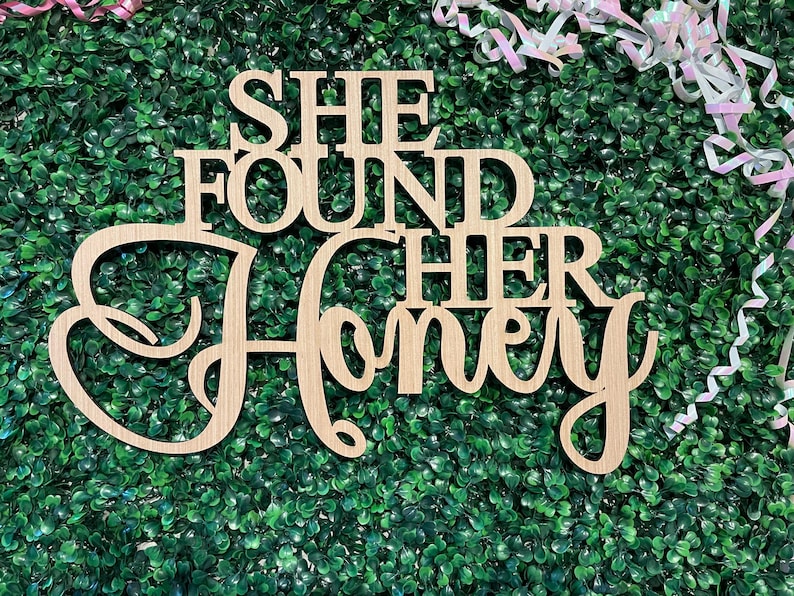 Peut inclure: Panneau en bois clair avec l'inscription "SHE FOUND HER HONEY" d&eacute;coup&eacute;e dans une police d&eacute;corative. Le panneau est plac&eacute; sur un fond de feuillage vert, avec des rubans roses et blancs en arri&egrave;re-plan.