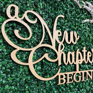 May include: Wooden sign with the words "A New Chapter Begins" in a cursive and block font. The sign is light brown and set against a backdrop of green foliage. Decorative ribbons are in the upper right corner.