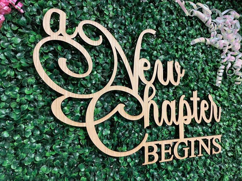 May include: Wooden sign with the words "A New Chapter Begins" in a cursive and block font. The sign is light brown and set against a backdrop of green foliage. Decorative ribbons are in the upper right corner.