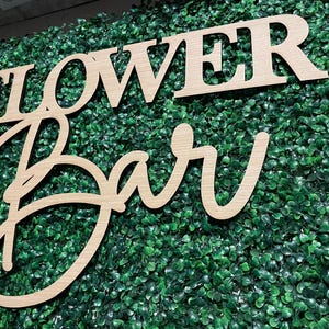 May include: Wooden sign with the words "Flower Bar" in a cursive font. The sign is placed on a green artificial grass backdrop.