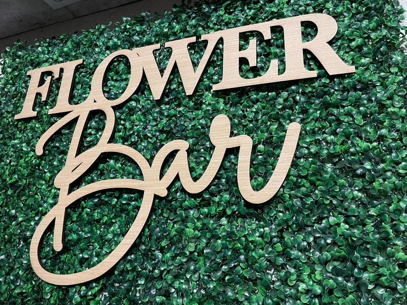 May include: Wooden sign with the words "Flower Bar" in a cursive font. The sign is placed on a green artificial grass backdrop.