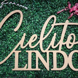 May include: Wooden sign with the words "Cielito Lindo" in a cursive font and block letters. The sign is made of light-colored wood and is set against a backdrop of green foliage. Decorative ribbons are in the background.